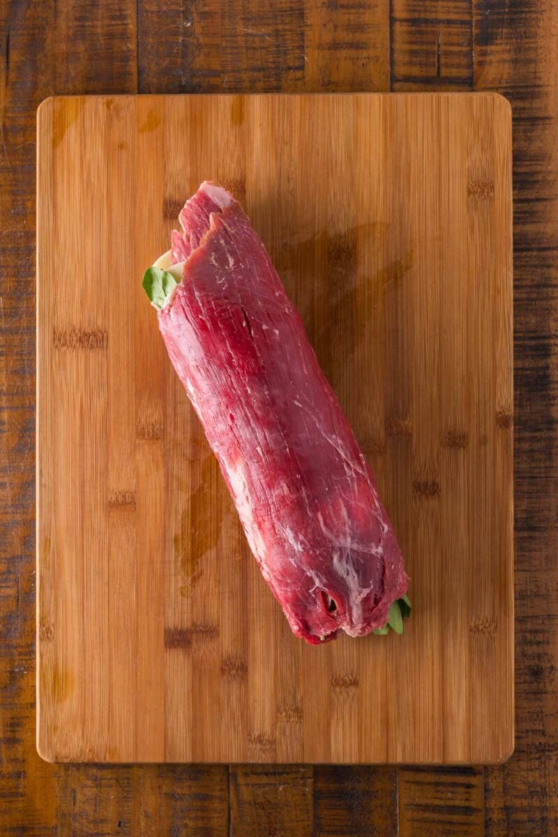 Raw flank steak rolled up with filling on a wooden cutting board.