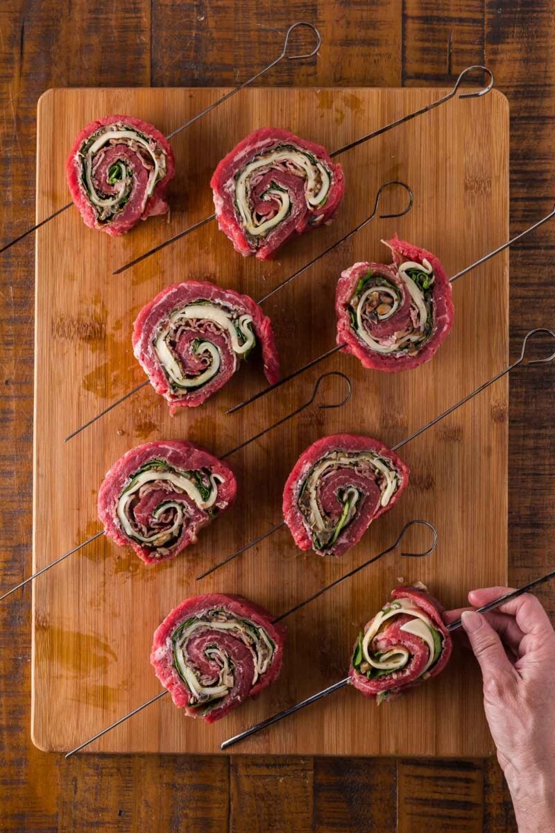 Eight raw beef pinwheels with spinach and cheese are arranged on metal skewers on a wooden cutting board; a hand is holding one skewer.