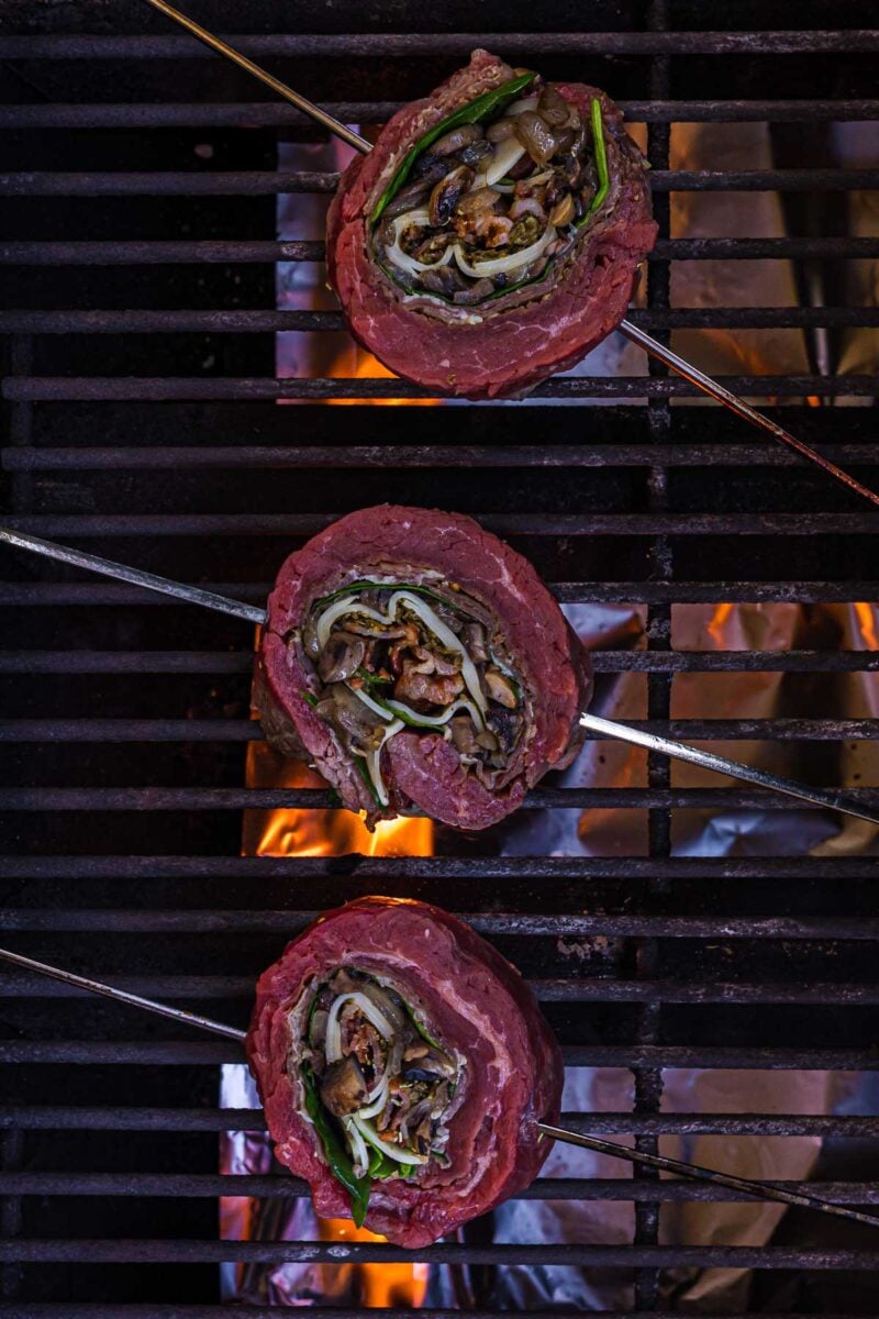 Three rolled and stuffed flank steak pinwheels on skewers are grilling over open flames on a barbecue grill.