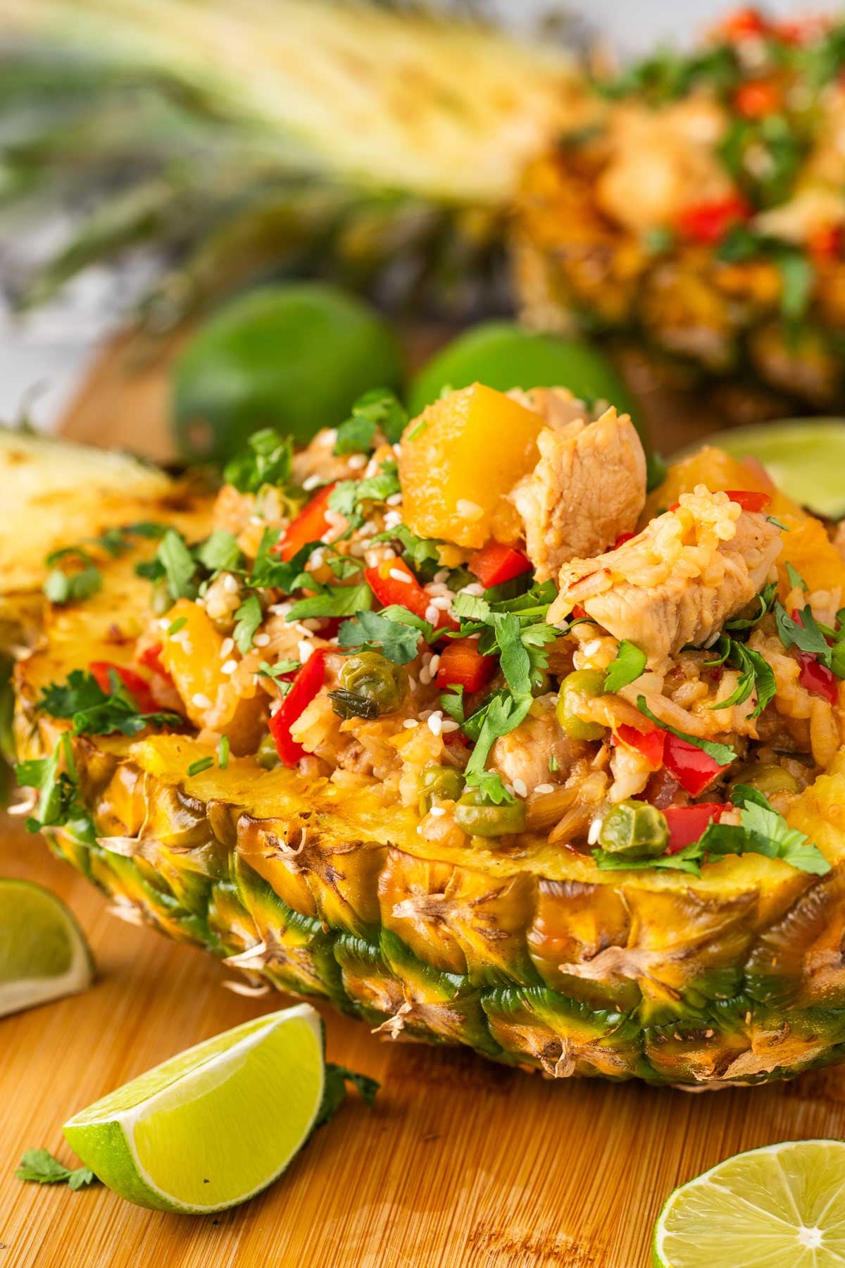 A halved pineapple filled with chicken, rice,sauted veggies, and herbs, garnished with chopped cilantro and served with lime wedges on a wooden surface.