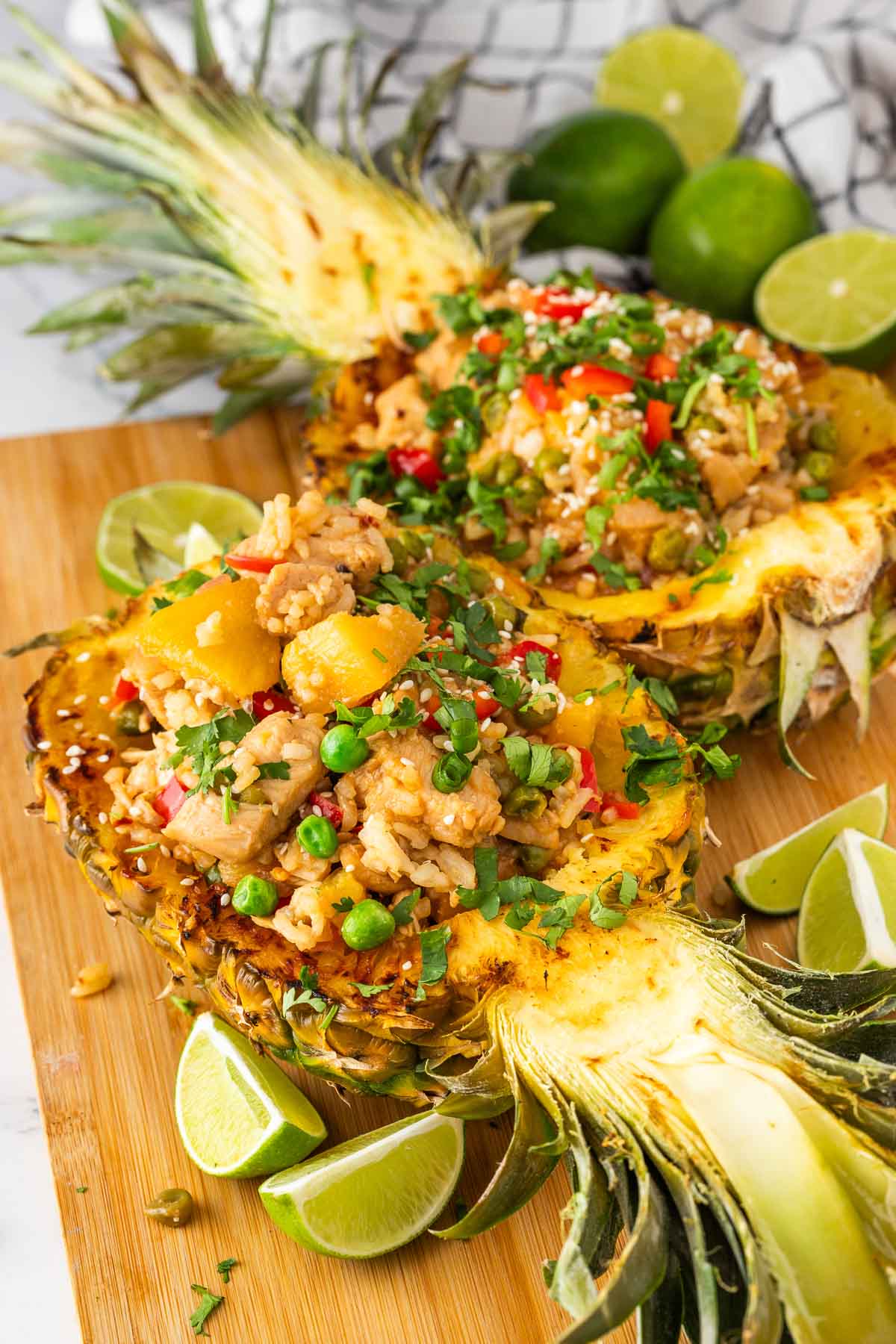 Two pineapple halves filled with fried rice, chicken, vegetables, and garnished with herbs and sesame seeds, placed on a wooden board with lime wedges and whole limes in the background.