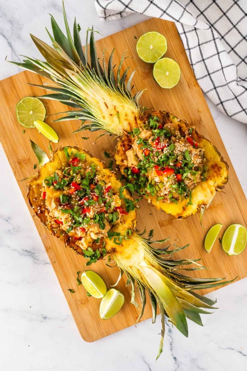 Two pineapple halves filled with rice, chicken, vegetables, and herbs, garnished with sesame seeds, served on a wooden board with lime wedges.