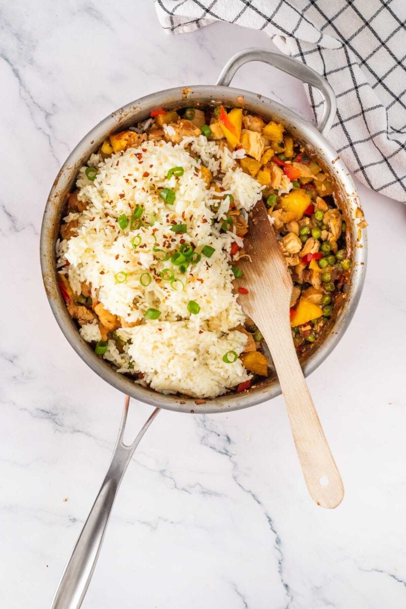 A stainless steel pan with cooked vegetables, diced chicken, and a mound of white rice topped with chopped green onions, on a marble surface with a checked kitchen towel.