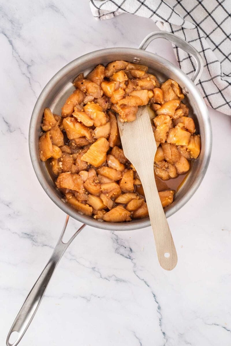 Chopped chicken pieces being cooked in a in sauce in a metal pan with a wooden spatula, on a marble countertop next to a checkered kitchen towel.