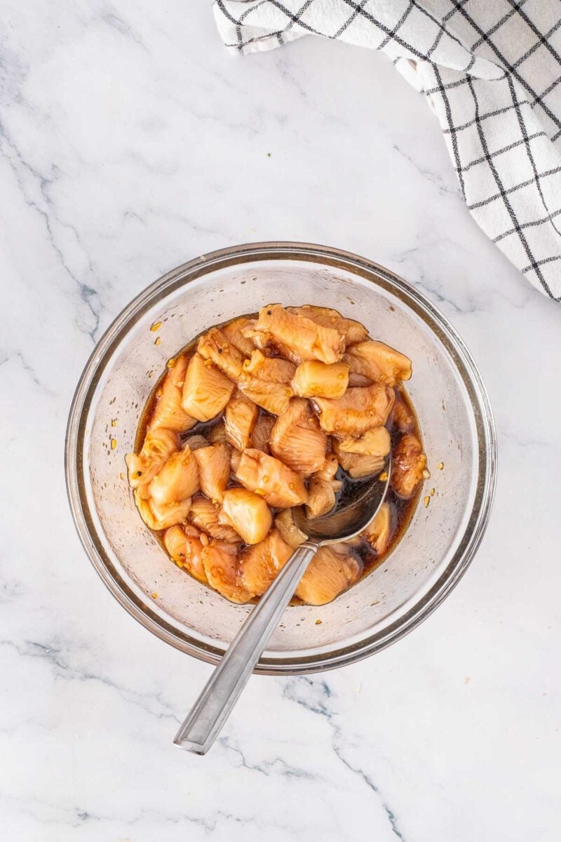 A glass bowl filled with marinated chunks of chicken and a metal spoon, placed on a white marble surface next to a checkered cloth.