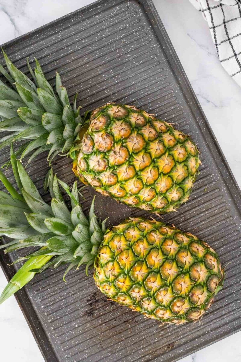 Two whole pineapples with green tops lie on grill pan.