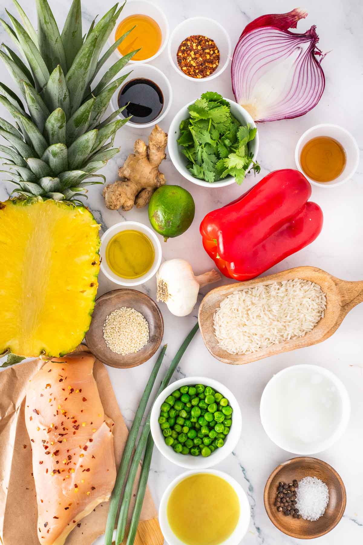 Assorted fresh ingredients including pineapple, chicken breast, rice, red bell pepper, peas, red onion, lime, cilantro, ginger, garlic, green onions, sauces, and seasonings on a white surface.