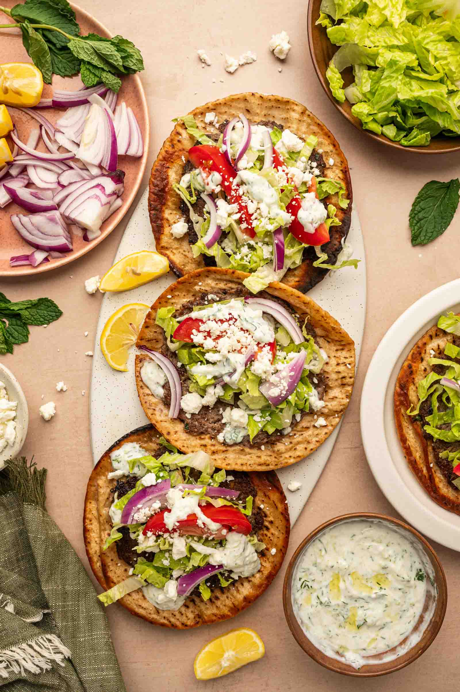 Three gyro smashburgers topped with lettuce, tomato, red onion, feta, and tzatziki sauce on a platter, surrounded by fresh vegetables, herbs, and lemon wedges.