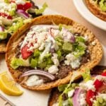 Gyro smashburgers topped with ground lamb, lettuce, tomatoes, red onion, feta cheese, and white sauce, served with lemon wedges.