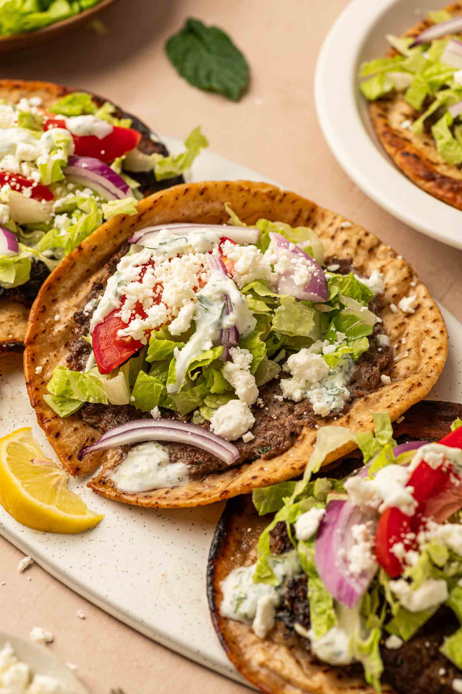 Gyro smashburgers topped with ground lamb, lettuce, tomatoes, red onion, feta cheese, and white sauce, served with lemon wedges.