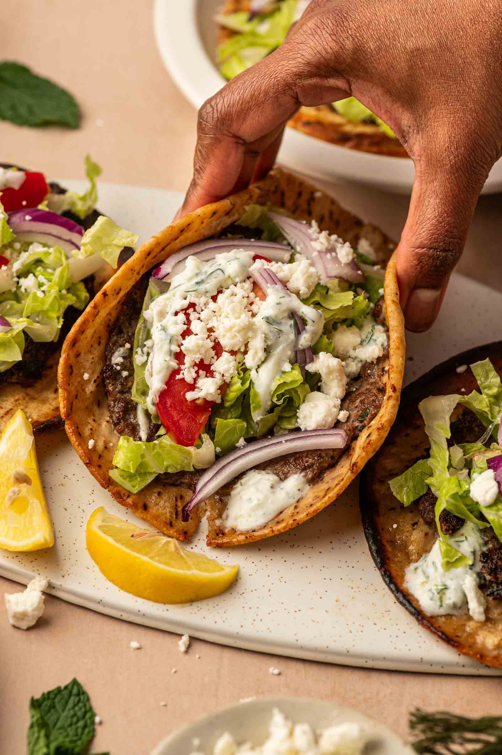 A hand holds a gyro smasshburger filled with ground lamb, lettuce, tomato, red onion, feta cheese, and a creamy white sauce, served on a white plate with lemon wedges.
