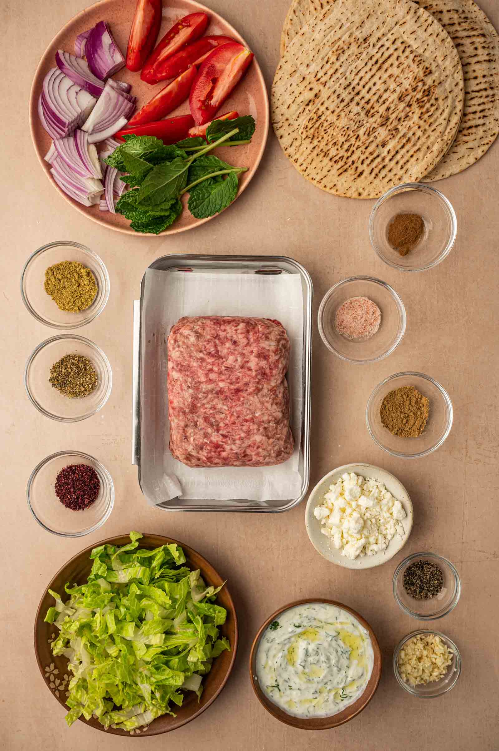 Ingredients for a gyro smashburger, including ground lmb, flatbread, vegetables, herbs, spices, feta cheese, tzatziki sauce, and chopped lettuce arranged on a light surface.