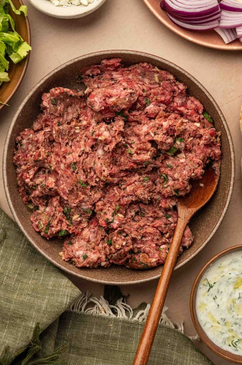 A bowl of seasoned raw ground lamb mixture with herbs, stirred with a wooden spoon, surrounded by sliced red onions, sauce, and greens.