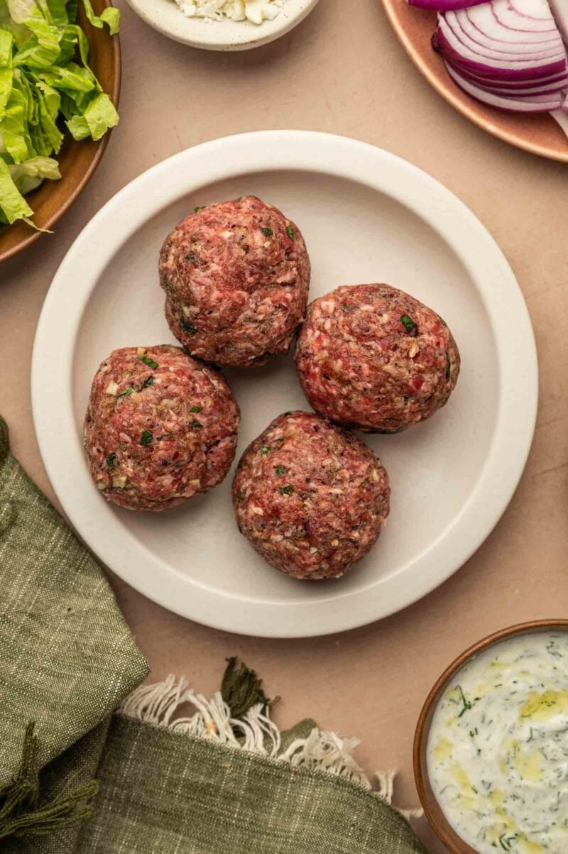 Four raw seasoned lamb smashburger balls are arranged on a round white plate, surrounded by dishes with lettuce, red onion slices, and a bowl of white sauce.