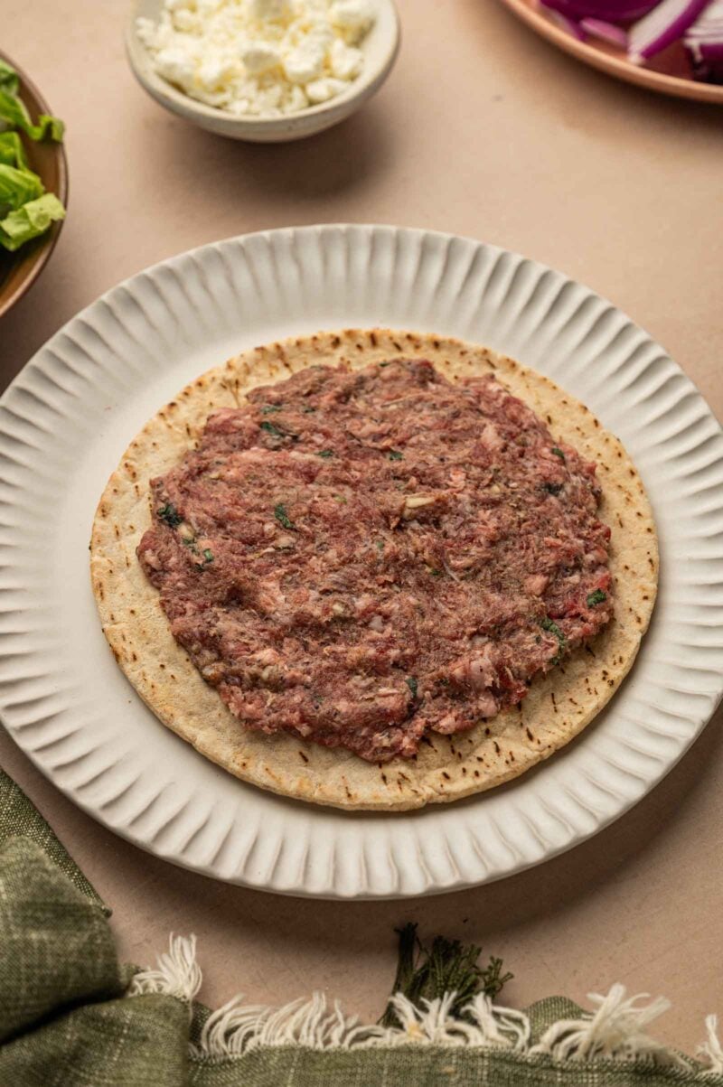 A flatbread topped with a raw minced lamb mixture sits on a white plate, with bowls of cheese, greens, and red onions nearby.