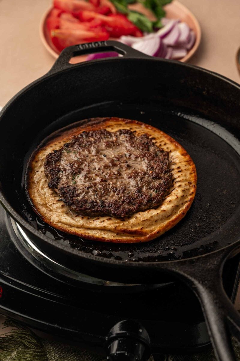 A lamb smashburger cooking on top of a flatbread in a black cast iron skillet, with sliced vegetables in the background.