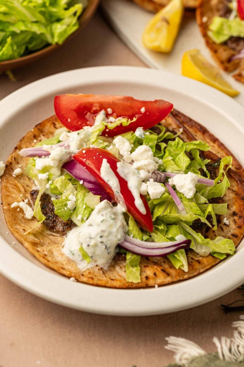 A gyro smashburger topped with lettuce, tomato wedges, red onion slices, crumbled feta cheese, and tzatziki sauce on a white plate.