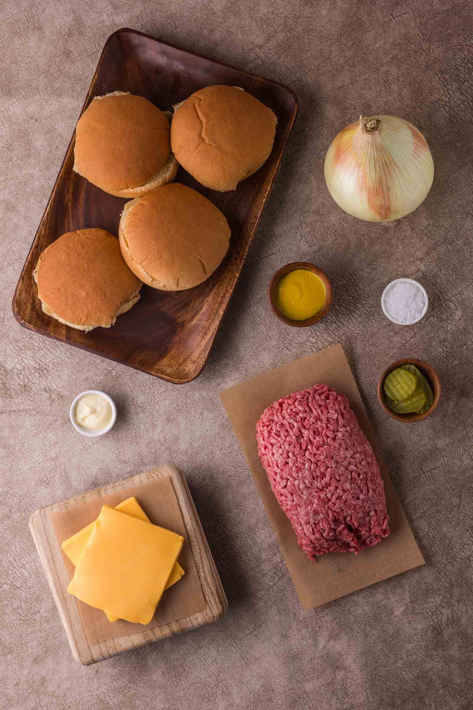 Top-down view of hamburger ingredients: four buns, a whole onion, ground beef, cheddar cheese slices, mayonnaise, pickle slices, salt, and oil arranged on a countertop.