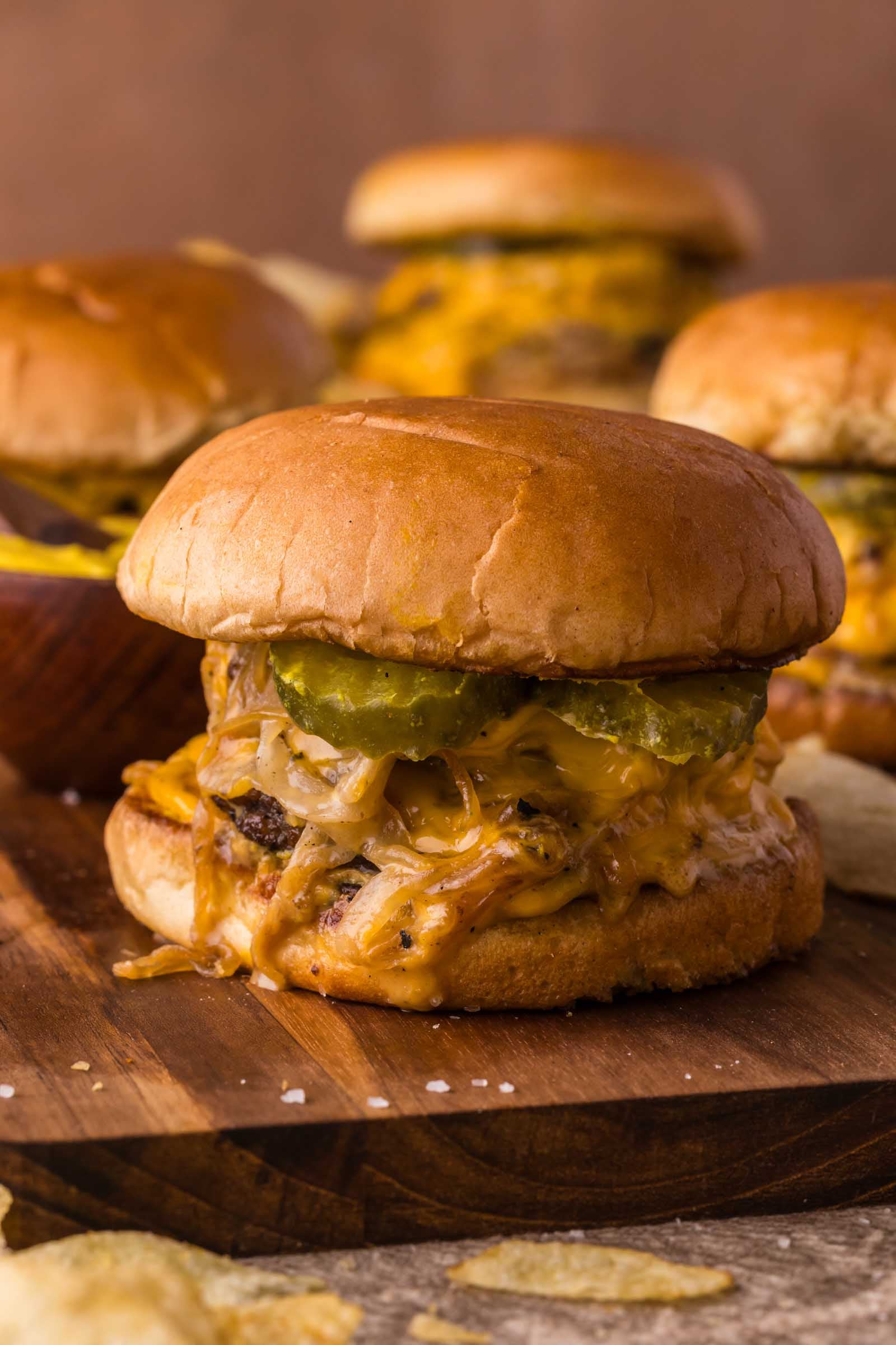 An Oklahoma burger with melted cheese, lots of onions, pickles, and a toasted bun sits on a wooden board, with more burgers and potato chips in the background.