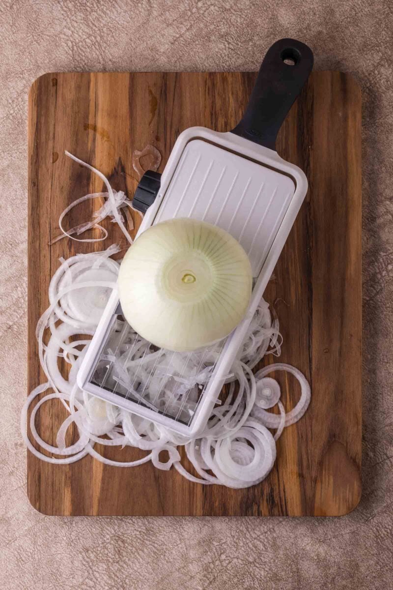 A white mandoline slicer with a whole onion and thinly sliced onion rings on a wooden cutting board.