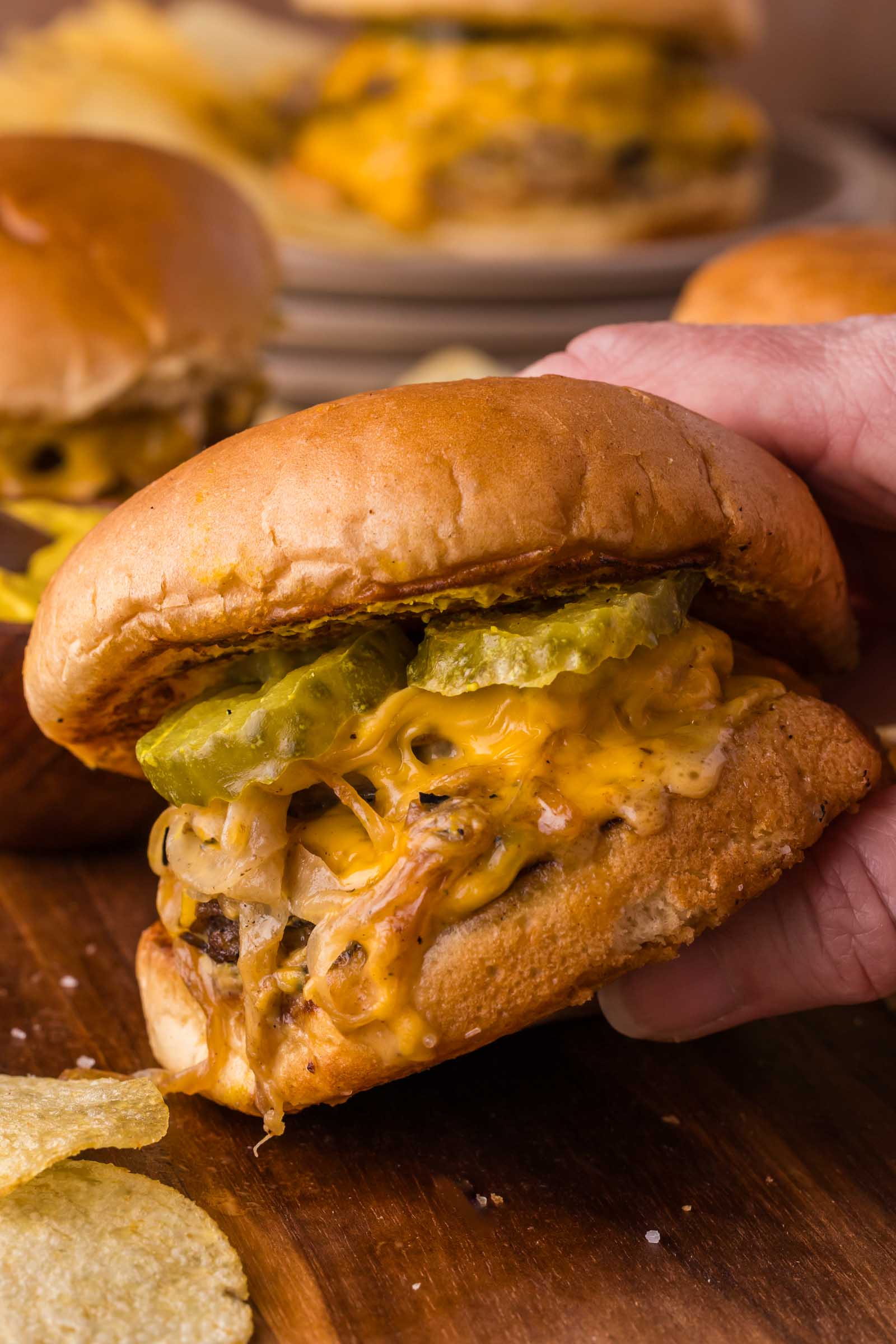 A hand holds an Oklahoma onion burger topped with pickles and onions on a wooden surface, with chips and other burgers in the background.