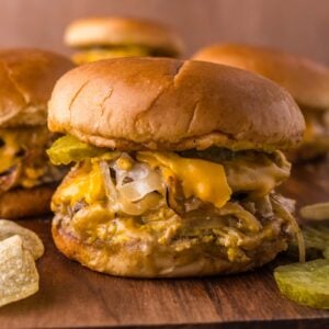 An Oklahoma onion burger with melted cheese, grilled onions, and pickles on a bun sits on a wooden board, with potato chips and more burgers in the background.