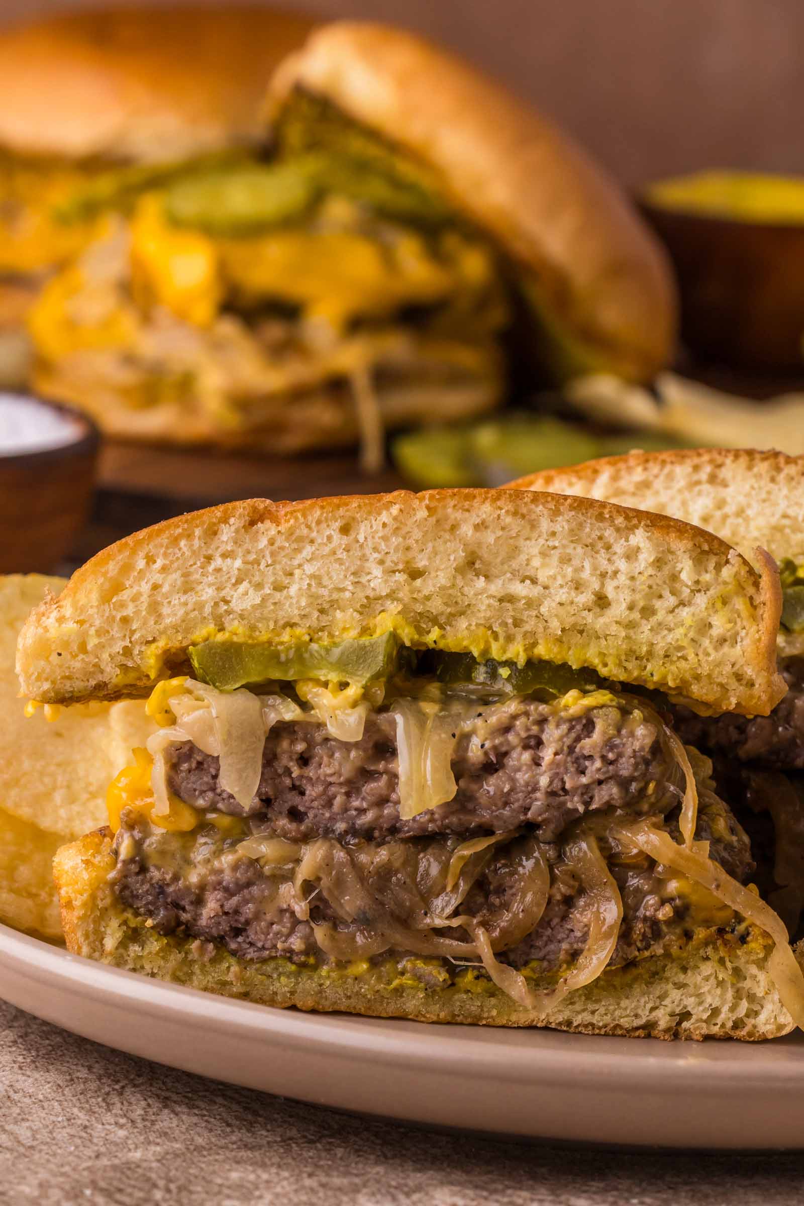 A close-up of a sliced Oklahoma onion burger with pickles, onions, and melted cheese on a white plate, with another burger in the background.