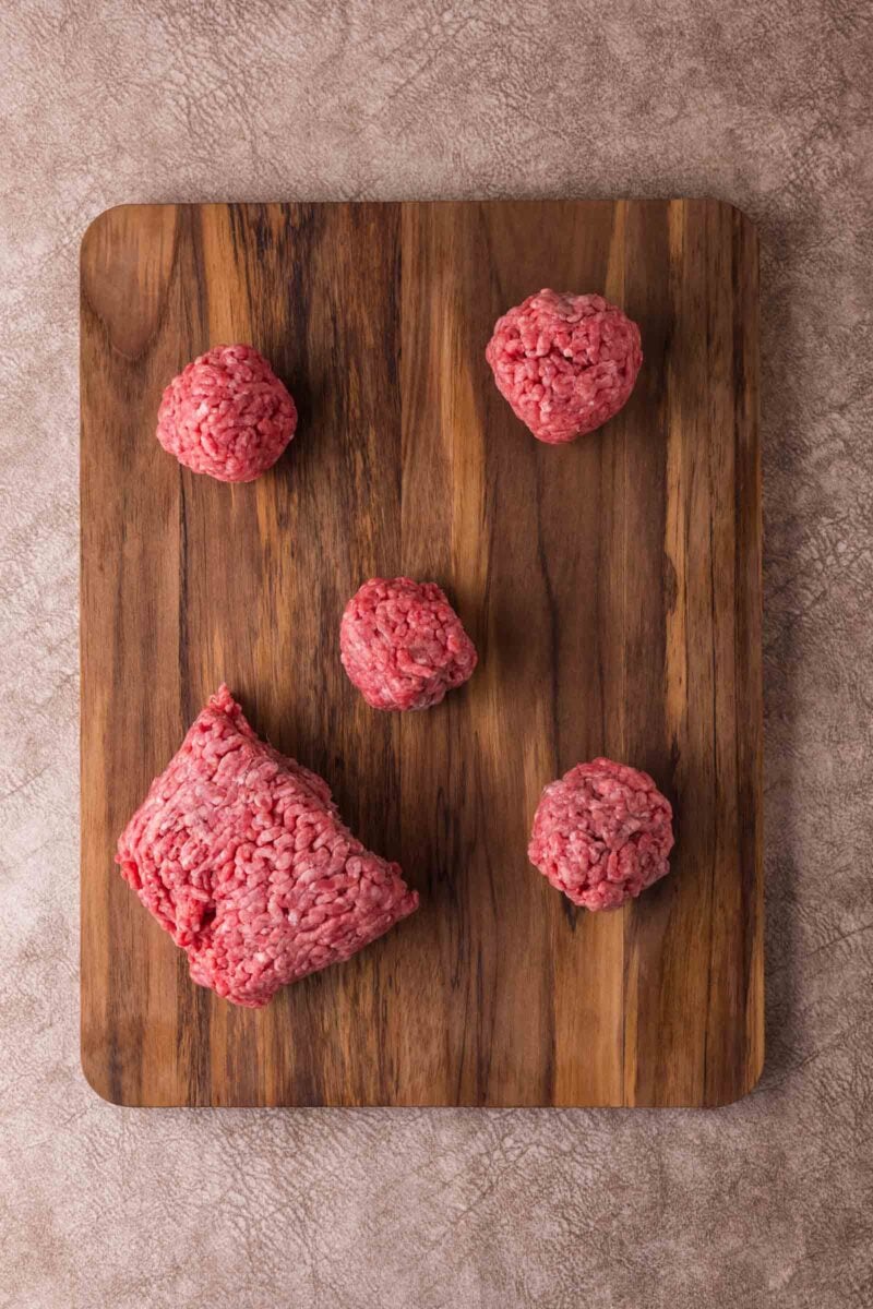 A wooden cutting board with a portion of raw ground beef being used to make round balls.
