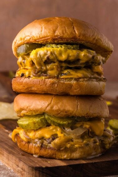 Two Oklahoma onion burgers stacked on top of each other, each with melted cheese, pickles, and grilled beef patties on toasted buns, placed on a wooden surface.