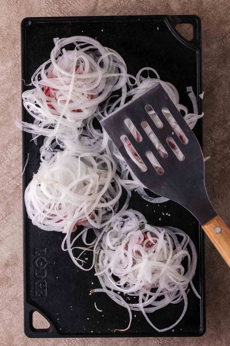 Four raw hamburger patties topped with thinly sliced onions on a black griddle, with a metal spatula resting on one patty.