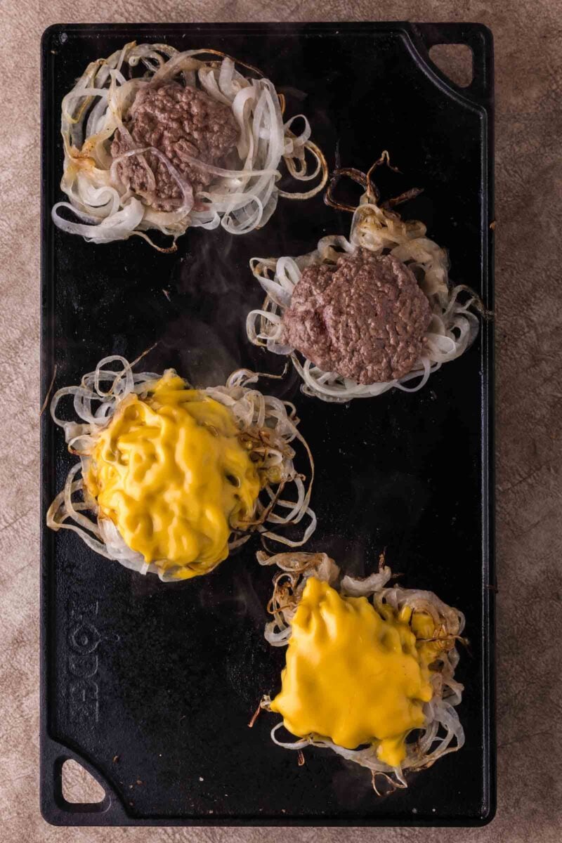 Four burger patties cooking on a griddle, each covering a mound of grilled onions; two patties are covered with melted yellow cheese.