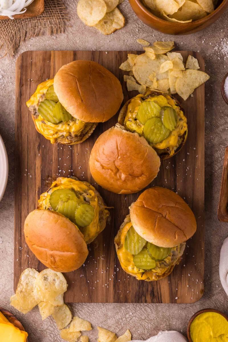 Four Oklahoma onion burgers topped with pickles are arranged on a wooden board, surrounded by potato chips.