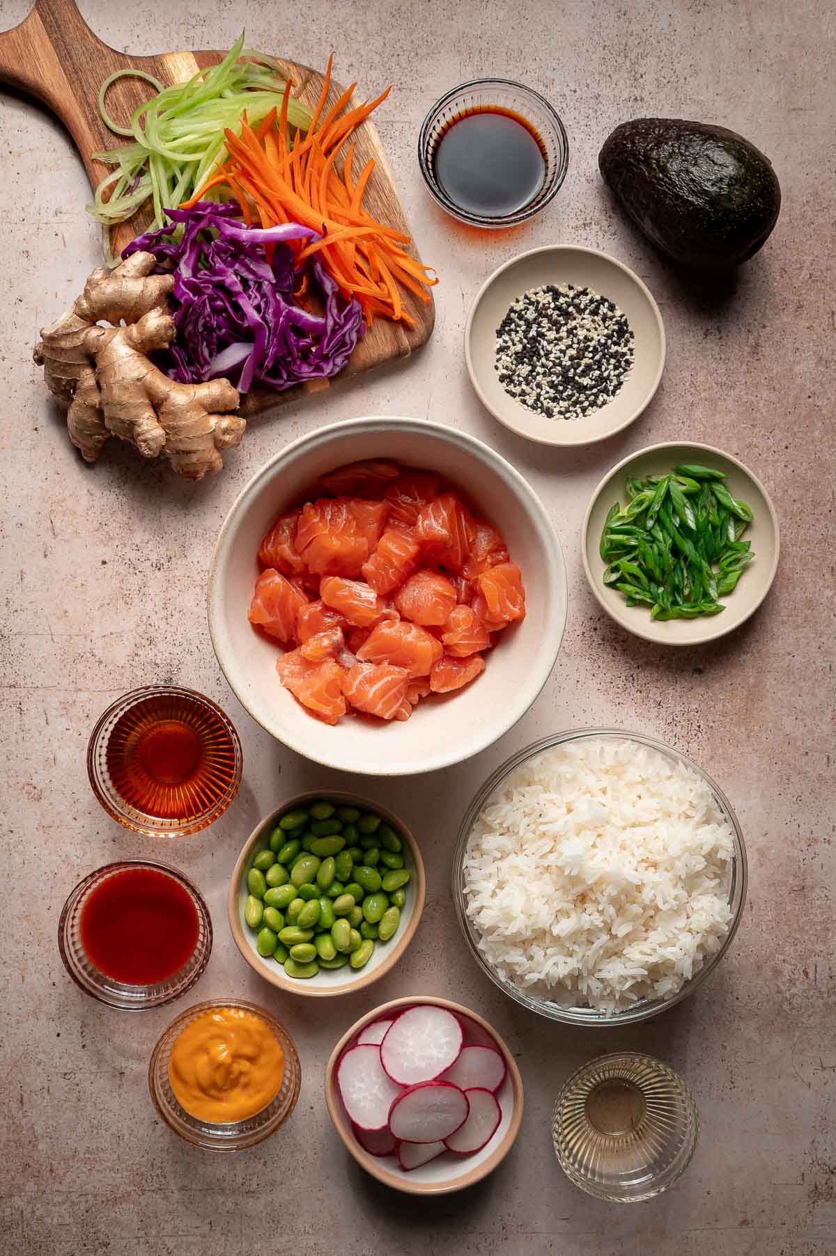 Overhead view of poke bowl ingredients, including diced raw salmon, rice, edamame, sliced vegetables, ginger, avocado, sauces, and sesame seeds arranged in bowls on a countertop.
