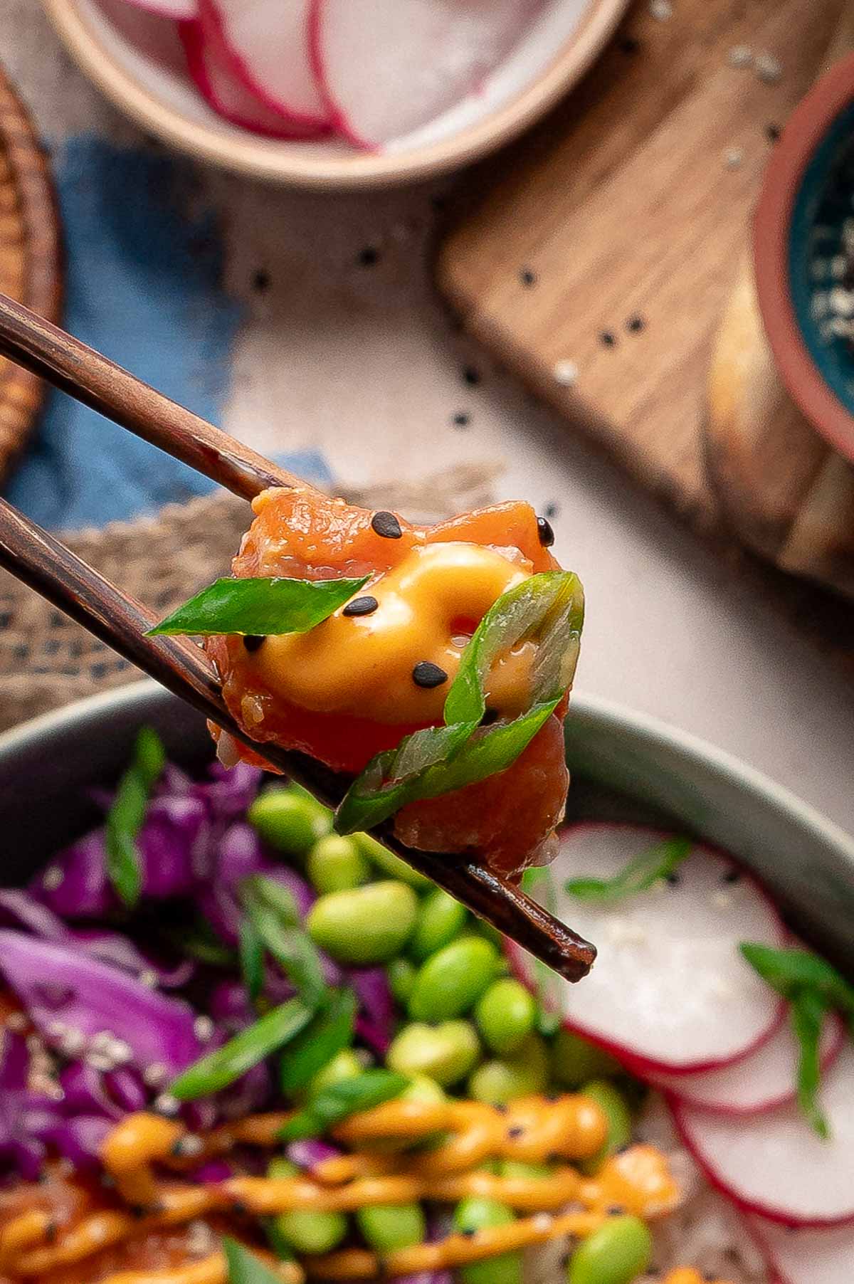 Close-up of chopsticks holding a piece of poke topped with sauce, green onion, and black sesame seeds above a bowl of assorted vegetables.