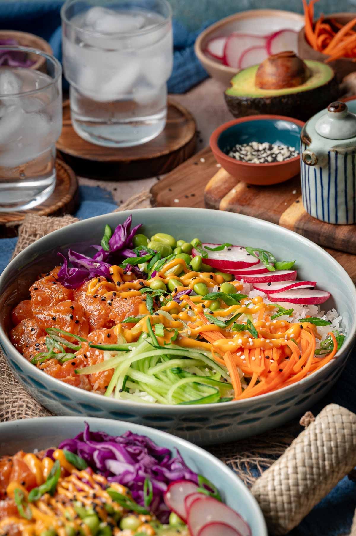 A bowl of poke with diced salmon, shredded carrots, cucumber, radish, cabbage, edamame, and a drizzle of sauce, served with glasses of ice water and avocado on the side.