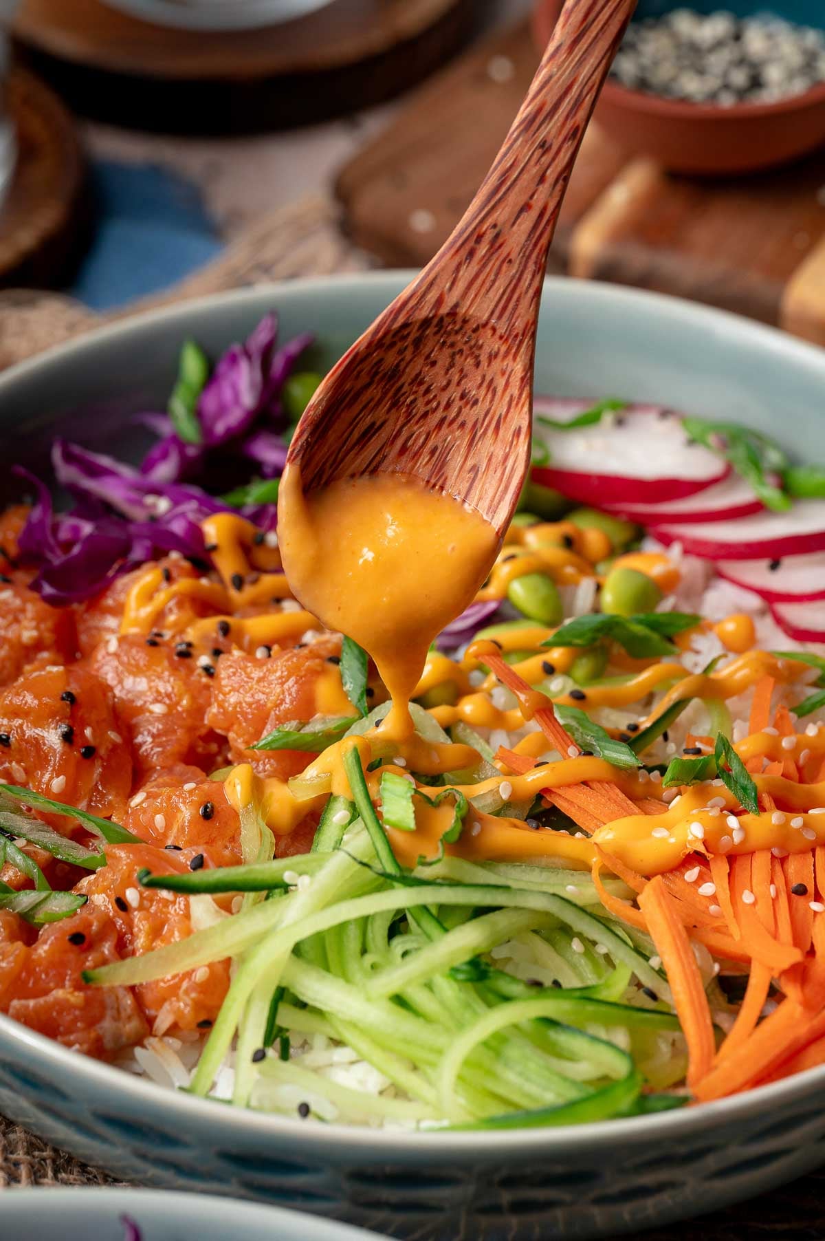 A wooden spoon drizzles orange sauce over a poke bowl of rice topped with raw salmon, shredded carrots, cucumber, radish, cabbage, edamame, and sesame seeds.