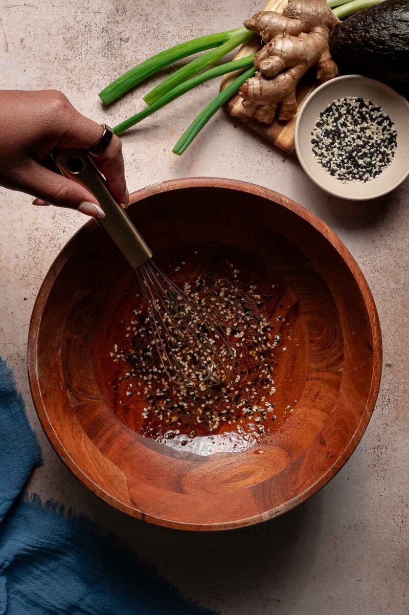 A hand uses a whisk to mix a marinade with sesame seeds in a wooden bowl. Green onions, ginger, and a small bowl of seeds are nearby on the counter.