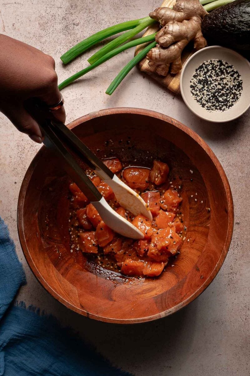A hand uses tongs to mix cubed marinated salmon in a wooden bowl; green onions, ginger, an avocado, and a bowl of sesame seeds are nearby.