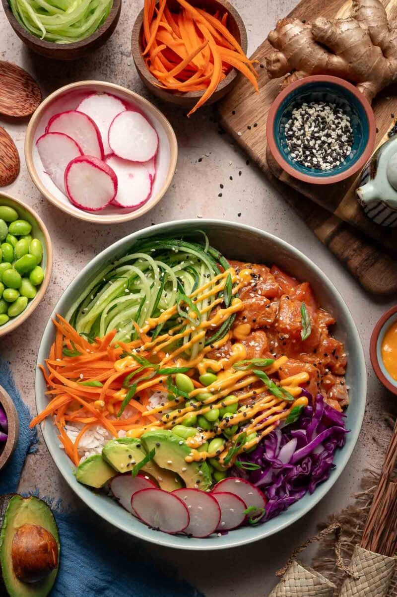 A colorful poke bowl with sliced cucumber, carrot, edamame, avocado, radish, cabbage, marinated salmon, and sauces, surrounded by small bowls of ingredients.