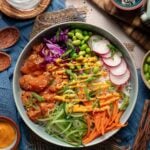 A poke bowl with diced fish, edamame, shredded carrot, cucumber, red cabbage, radish slices, rice, green onions, and sauce, surrounded by small bowls of ingredients.