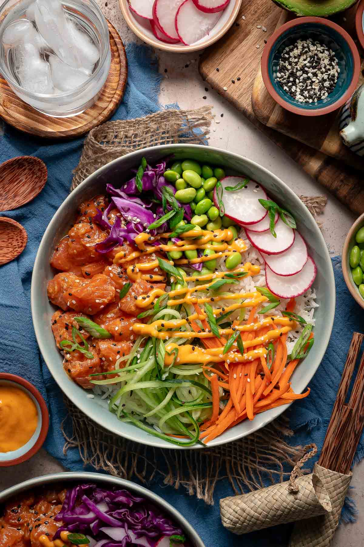 A poke bowl with diced fish, edamame, shredded carrot, cucumber, red cabbage, radish slices, rice, green onions, and sauce, surrounded by small bowls of ingredients.