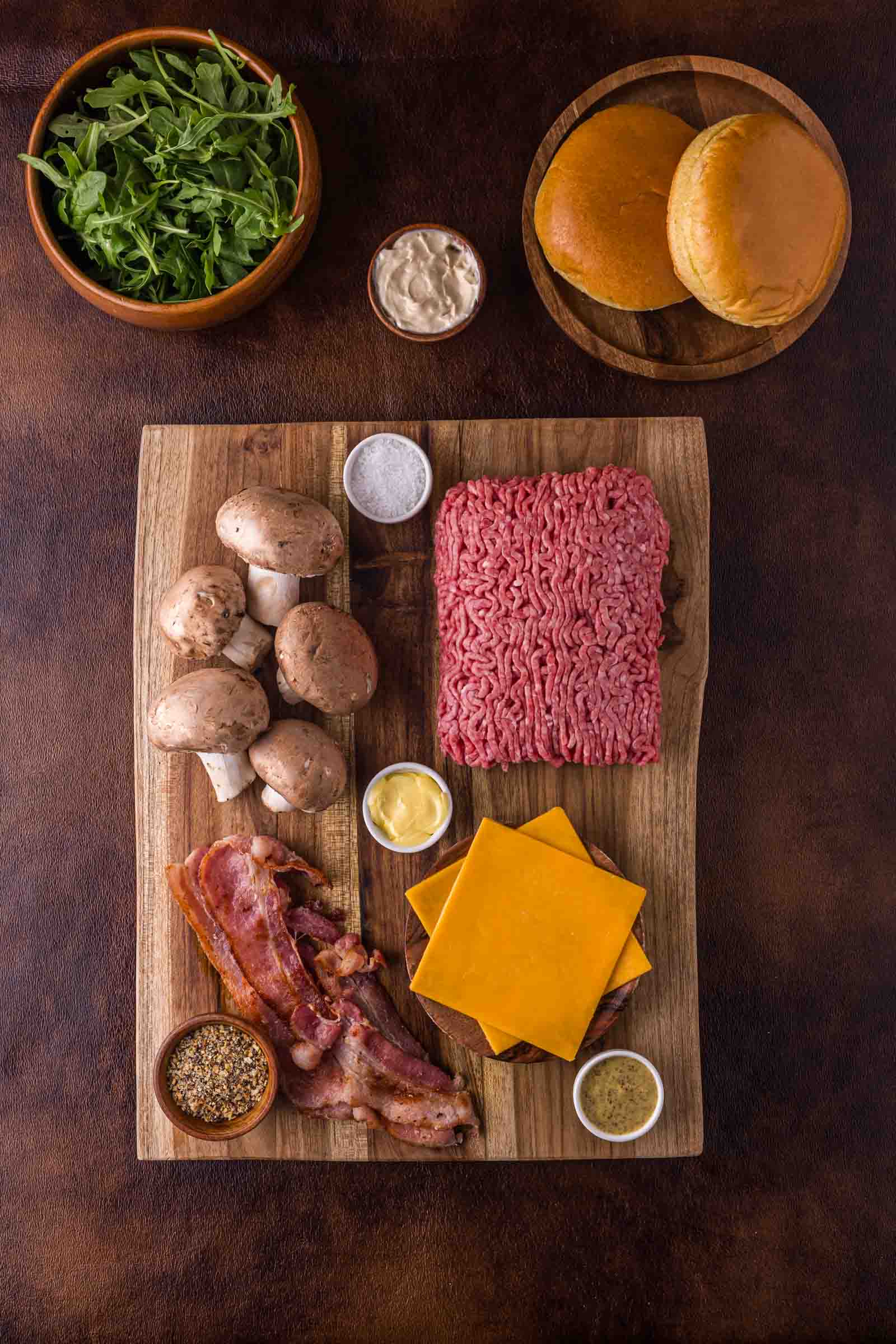 A wooden board with raw ground beef, cheddar cheese slices, bacon strips, mushrooms, condiments, and seasonings. Arugula, burger buns, and sauce are placed nearby on the table.