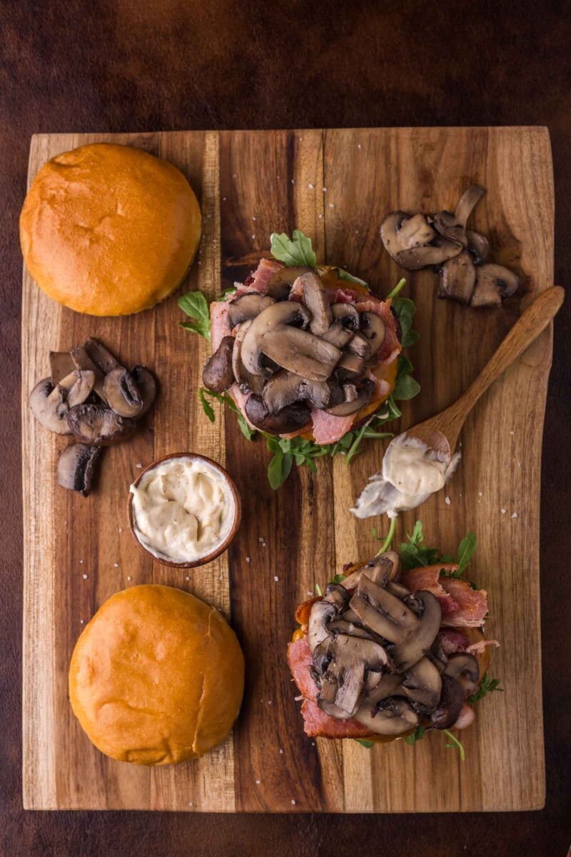 A wooden board with two open burgers topped with arugula, bacon, and sautéed mushrooms, a small dish of mayonnaise, a spoon, and two sandwich buns.