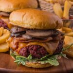 Close-up of a gourmet smoked steakhouse burger with sautéed mushrooms, bacon, cheddar cheese, and greens on a brioche bun, served with thick-cut fries. Another burger and fries are in the background.