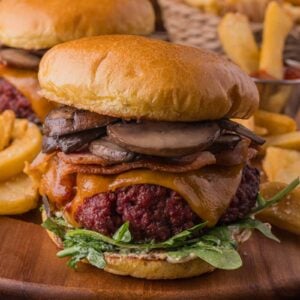 Close-up of a gourmet smoked steakhouse burger with sautéed mushrooms, bacon, cheddar cheese, and greens on a brioche bun, served with thick-cut fries. Another burger and fries are in the background.