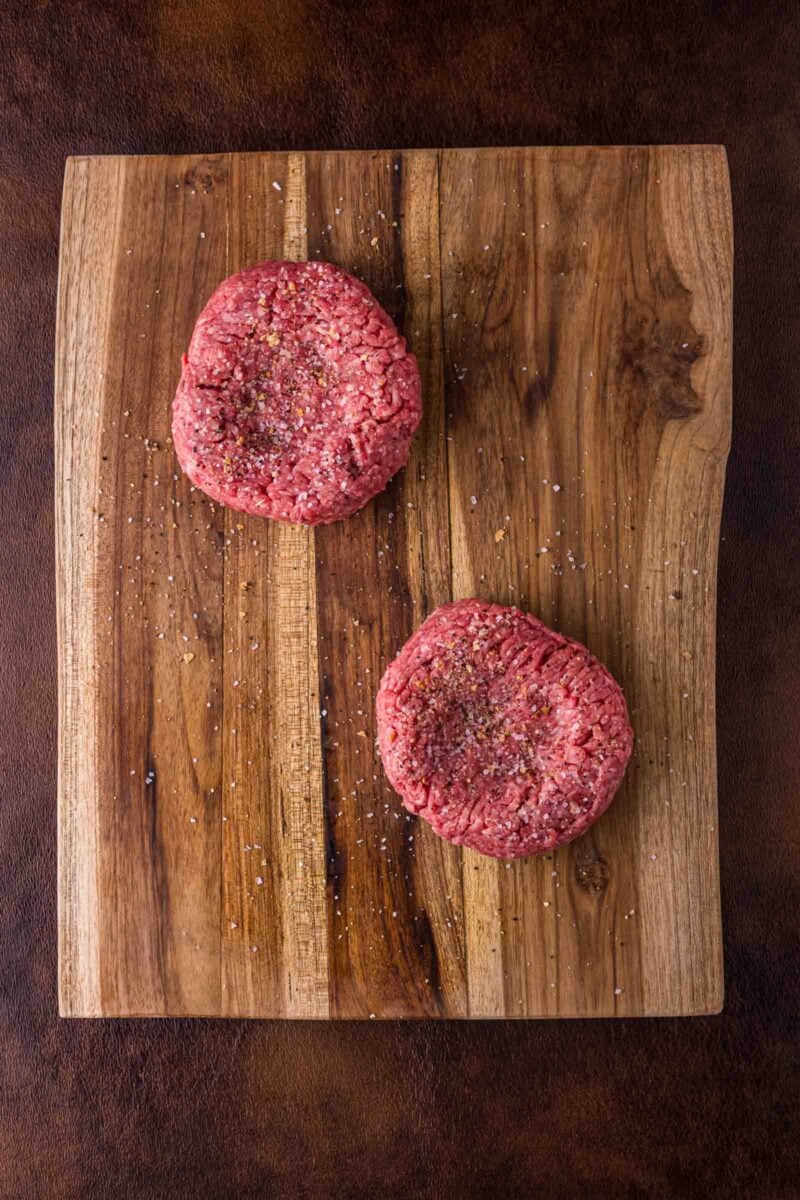 Two raw beef burger patties sprinkled with salt and pepper sit on a wooden cutting board.