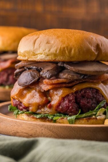 A close-up of a smoked steakhouse burger with lettuce, cheddar cheese, bacon, sautéed mushrooms, and a beef patty on a wooden plate.