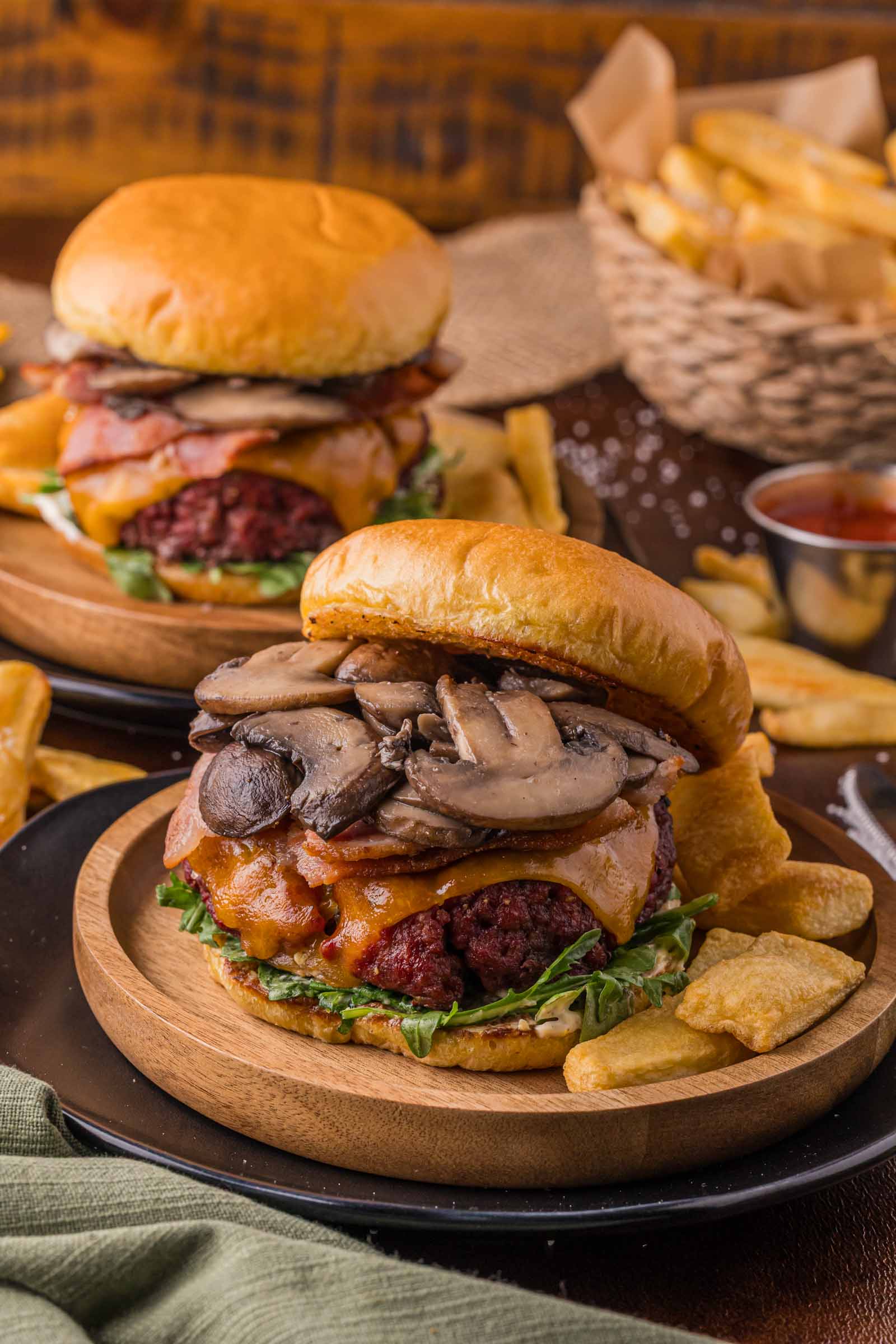 Two smoked steakhouse burgers topped with mushrooms, cheese, lettuce, and bacon are served on wooden plates with thick-cut fries; a basket of fries and ketchup are in the background.