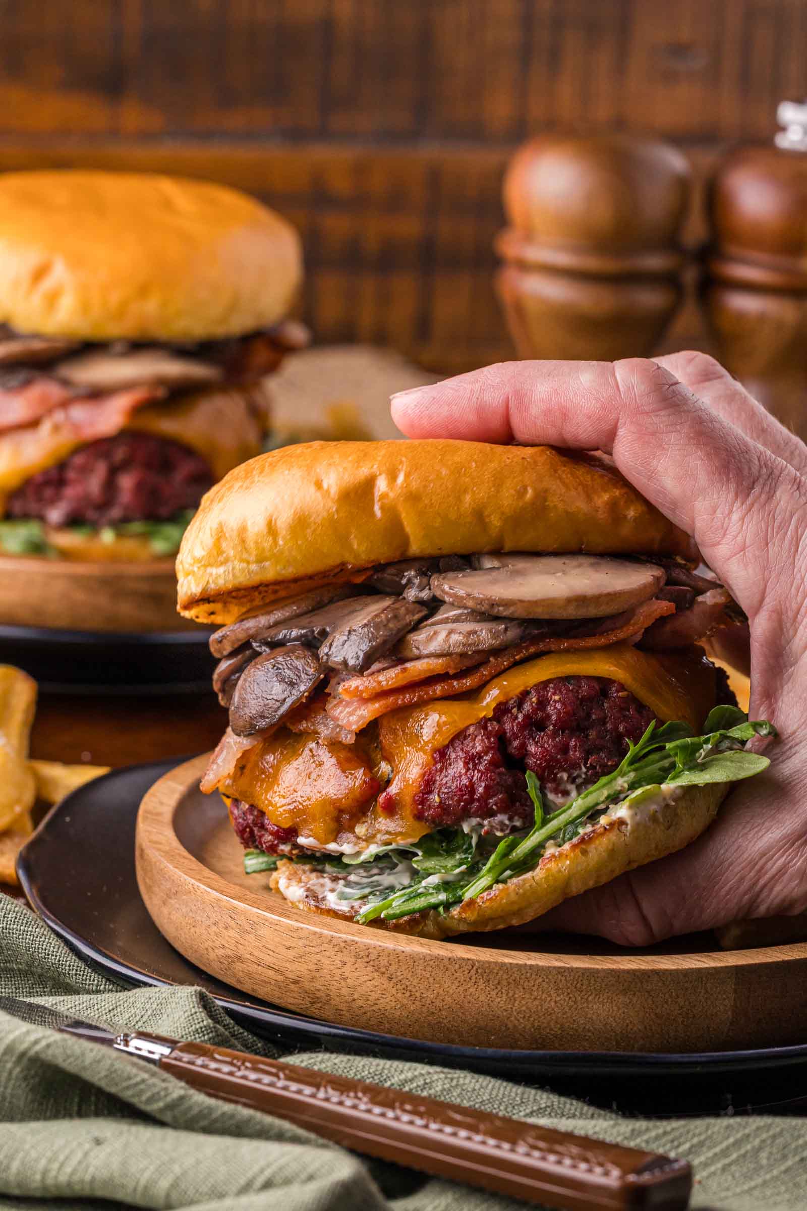 A hand holds a large smoked steakhouse burger with lettuce, cheddar cheese, bacon, mushrooms, and sauce in a toasted bun, served on a wooden plate with another burger and fries in the background.