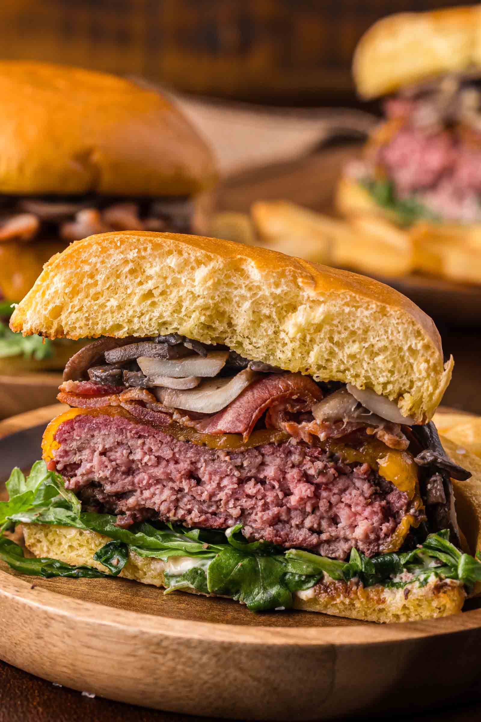 A close-up of smoked steakhouse burger cut in half to show the red center with lettuce, bacon, mushrooms, onions, and a beef patty on a wooden plate.